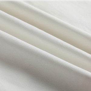 May include: A solid white fabric featuring a distinct diagonal twill weave pattern. The material is arranged in soft, flowing folds that create subtle shadows and highlights across the surface, emphasizing the texture of the woven textile.