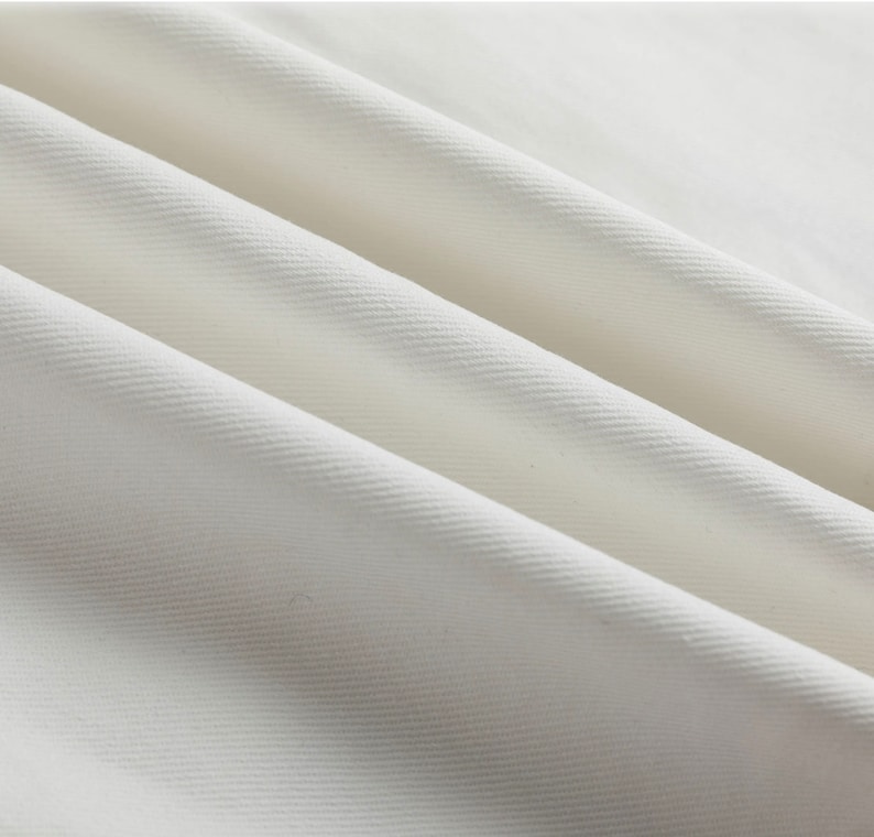 May include: A solid white fabric featuring a distinct diagonal twill weave pattern. The material is arranged in soft, flowing folds that create subtle shadows and highlights across the surface, emphasizing the texture of the woven textile.