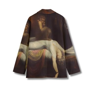 May include: A long-sleeve blazer featuring a dark, dramatic art print of a figure in a white garment reclining on a bed with a dark, shadowy creature looming above. The jacket has a structured collar, long sleeves, and a straight hemline.