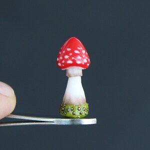 Könnte beinhalten: Ein Miniatur-Pilz in Rot und Weiß mit grünen und braunen Details wird von einer Pinzette vor einem grauen Hintergrund gehalten.