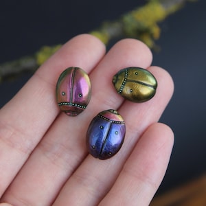 May include: Three iridescent beetles, one purple, one blue, and one gold, are displayed on a hand. The beetles are small and have a shiny finish.