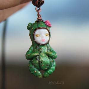 Puede incluir: Un colgante que muestra una pequeña figura de rostro pálido con un traje verde texturizado similar a un cactus, con puntos dorados y una pequeña flor rosa en la cabeza. La figura cuelga de una anilla de metal cobrizo en un cordón negro.