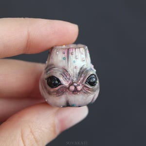 May include: A small, hand-sculpted figurine of a creature with large black eyes, a pink nose, and a light gray complexion. The figurine is adorned with small, colorful embellishments and is held in a person's hand.