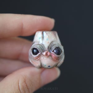 May include: A detailed miniature sculpture of an alien-like creature's head. The sculpture has large, dark eyes with sparkling details, a pink nose, and pale, textured skin with subtle colour variations and small, colourful accents.