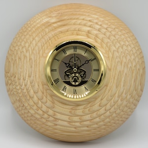 Könnte beinhalten: Eine runde Holzuhr mit hellem Holzmaserungsmuster. Das Zifferblatt hat eine goldfarbene Lünette mit römischen Ziffern und ein sichtbares mechanisches Uhrwerk. Ein dekorativer Artikel.