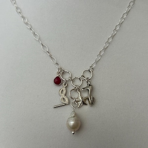 May include: A silver necklace with multiple charms. The charms include a red gemstone, a masquerade mask, a starfish, a deer, and a large pearl. The chain is made of oval links.