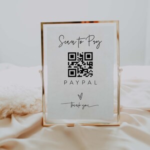 Puede incluir: Un cartel blanco con un marco dorado en un soporte. El cartel dice "Scan to Pay" con un código QR en blanco y negro y el texto "PAYPAL" debajo. Un símbolo de corazón y el texto "Gracias" están debajo del logotipo de PayPal. El texto "Calligraphy Co." está en la parte inferior de la imagen.