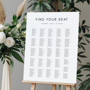 Könnte beinhalten: Eine weiße Sitzordnung mit dem Text „FIND YOUR SEAT“ und Namen von Gästen in Spalten. Das Schild wird auf einer hölzernen Staffelei präsentiert. Blumen sind im Hintergrund zu sehen.