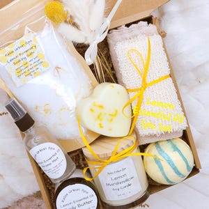 May include: A gift box filled with spa products, including a heart-shaped soap, a candle, bath salts, a loofah, and a bath bomb. The box contains a bottle of aroma mist and body butter. The products are lemon-scented and tied with yellow ribbons.