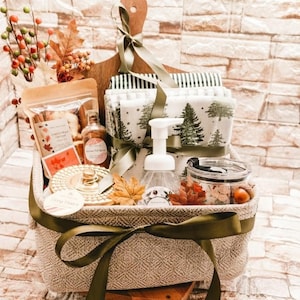 May include: A woven basket containing a wooden cutting board, folded white fabric with green tree patterns, a foaming soap dispenser, a glass jar, a pouch of dried goods, and decorative autumn leaves tied with a green ribbon.