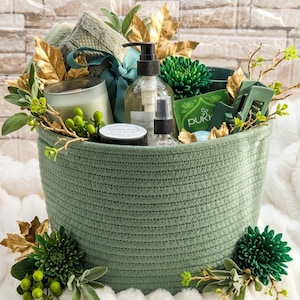 May include: A sage green woven rope basket filled with self-care items including a glass candle, a pump bottle, a small spray bottle, a Pukka tea packet, a green hair clip, a folded green towel, and decorative gold leaves and green flowers.
