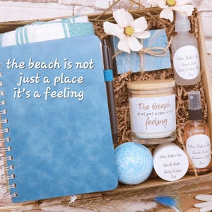 May include: A gift box containing a blue spiral-bound notebook with the text 'the beach is not just a place it's a feeling', a black pen, a candle with the same text, a blue bath bomb, body butter, and two spray bottles labeled 'Relax Urban Spa'.