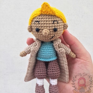 May include: Crocheted amigurumi doll with brown hair, a yellow spiky top, a brown coat, a blue shirt, brown trousers, and white socks. The doll is holding its hands in front of it.
