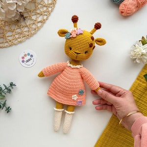 May include: A crocheted giraffe doll wearing a pink dress with white crocheted socks and a flower crown. The doll is sitting on a white surface with yarn balls and flowers in the background.