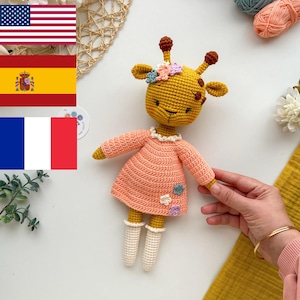 Può includere: Un giocattolo di giraffa all'uncinetto che indossa un vestito rosa con calzini bianchi all'uncinetto e una corona di fiori. La girafa è seduta su una superficie bianca con gomitoli di lana e un cesto intrecciato sullo sfondo.