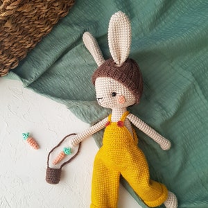 May include: A handmade crochet bunny doll wearing yellow overalls and a brown hat. The bunny has long ears, a small pink nose, and is holding a small brown bag with two crochet carrots. The doll is approximately 12 inches tall.
