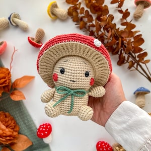 May include: A crocheted mushroom doll with a red cap and white spots, beige body, and a green bow. The doll has a smiling face with rosy cheeks. Other crocheted mushrooms in various colors are scattered around.