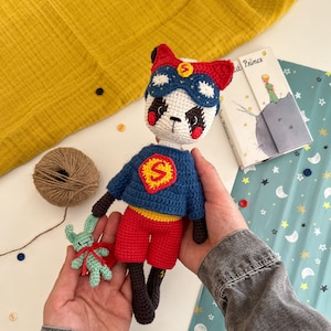 Può includere: Un panda all'uncinetto che indossa un costume da supereroe blu con una 'S' rossa sul petto e una maschera rossa. Il panda tiene un piccolo coniglietto verde all'uncinetto nella sua mano.