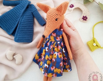 May include: A handmade crocheted fox doll, featuring an orange body and a blue floral dress. Accessories include a blue coat, white booties, a yellow purse, and a floral collar. A charming, handcrafted item for children.
