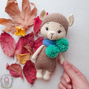 May include: A crocheted llama toy wearing a brown hat, blue scarf, and green pom-poms. The llama is sitting on a white background with autumn leaves.