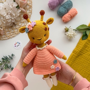 May include: A crocheted giraffe toy wearing a pink dress with flower accents. The giraffe is yellow with brown spots and has a white face.