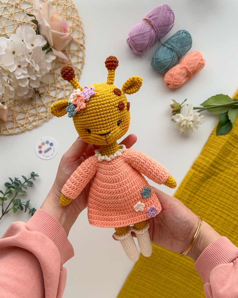 May include: A crocheted giraffe toy wearing a pink dress with flower accents. The giraffe is yellow with brown spots and has a white face.