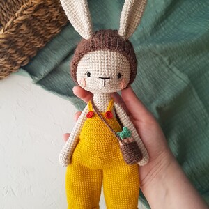 May include: Crocheted bunny doll with a cream-colored body, brown hat, and yellow overalls. The doll has long ears, a small brown bag, and red buttons. The doll is approximately 10 inches tall.