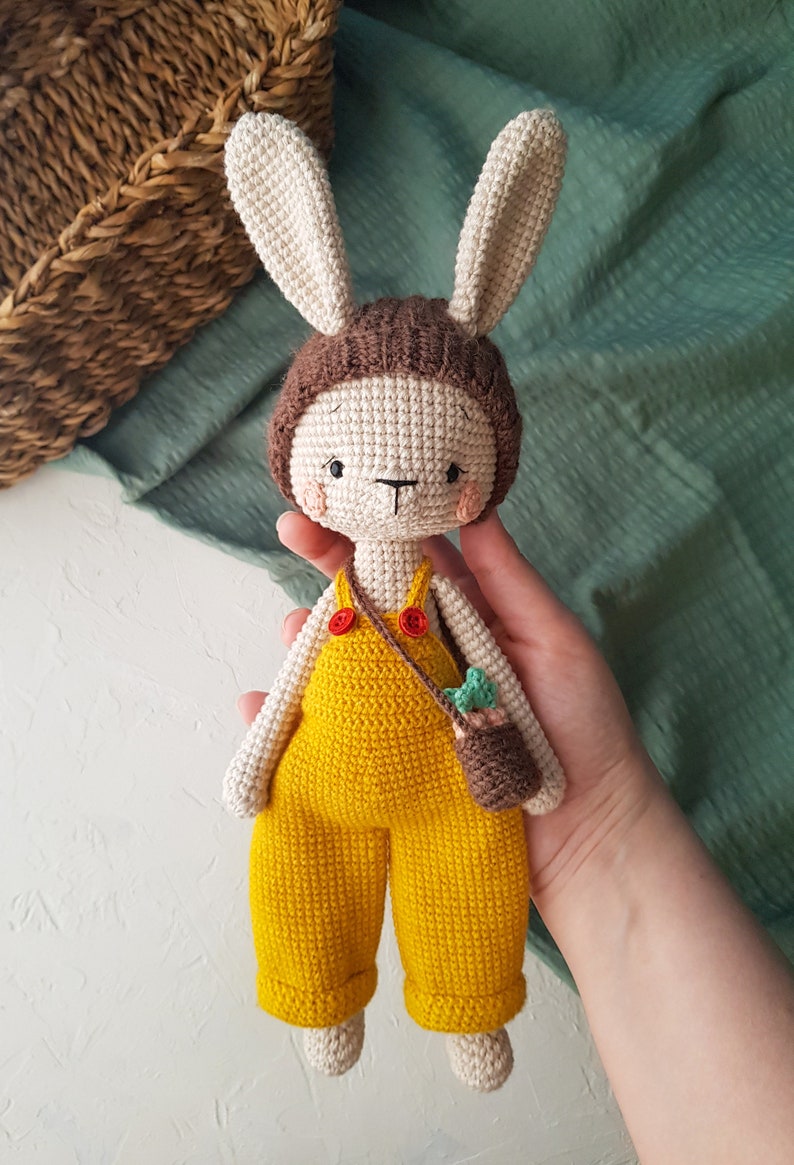 May include: Crocheted bunny doll with a cream-colored body, brown hat, and yellow overalls. The doll has long ears, a small brown bag, and red buttons. The doll is approximately 10 inches tall.