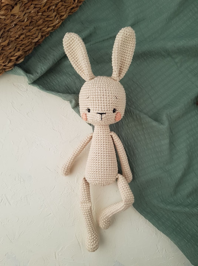 May include: A handmade, beige crochet bunny doll with long ears, a small black nose, and rosy cheeks. The bunny has a simple, endearing design, with arms and legs attached to the body. The doll is set against a white and green backdrop.