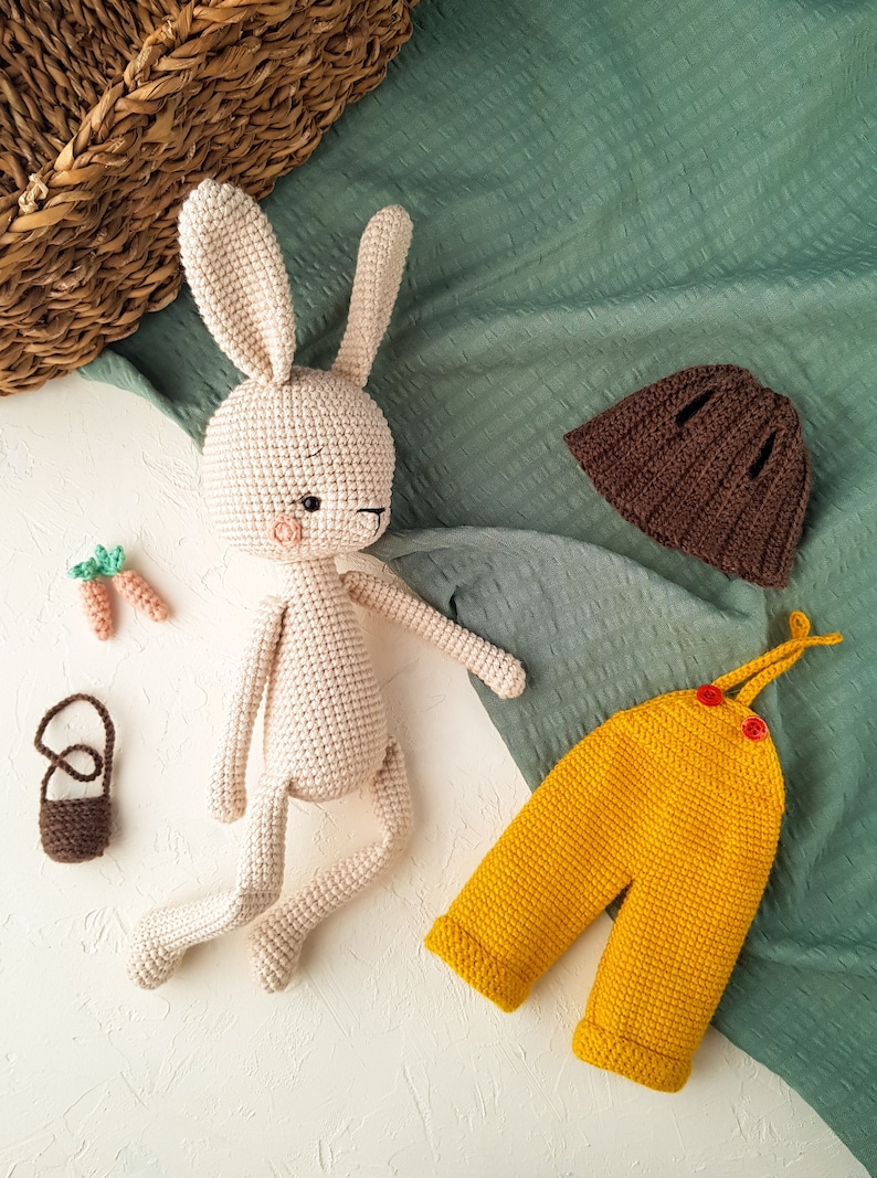 May include: A crocheted bunny doll with a brown hat and yellow overalls. The bunny is beige with a pink blush and black eyes. Two small crocheted carrots and a brown purse are also present.