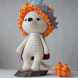 May include: A crocheted lion toy wearing a crown. The lion is white with orange and red accents. The crown is gray with orange accents. The lion has a long tail.
