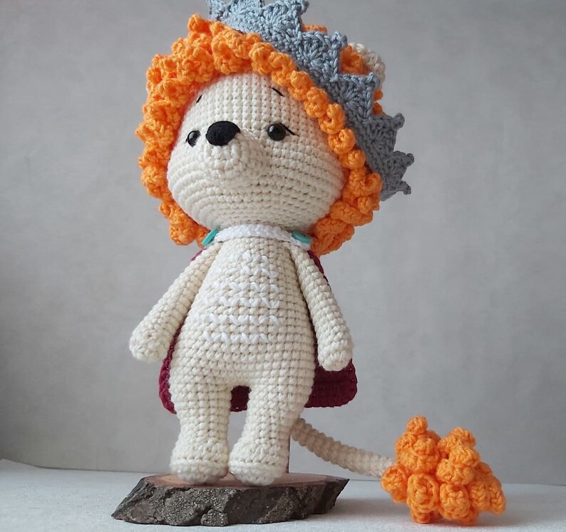 May include: A crocheted lion toy wearing a crown. The lion is white with orange and red accents. The crown is gray with orange accents. The lion has a long tail.