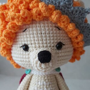 May include: A crocheted stuffed animal with a white body, orange hair, and a gray crown. The animal has black eyes and a black nose.