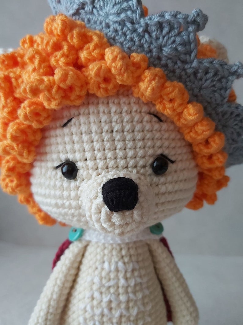 May include: A crocheted stuffed animal with a white body, orange hair, and a gray crown. The animal has black eyes and a black nose.