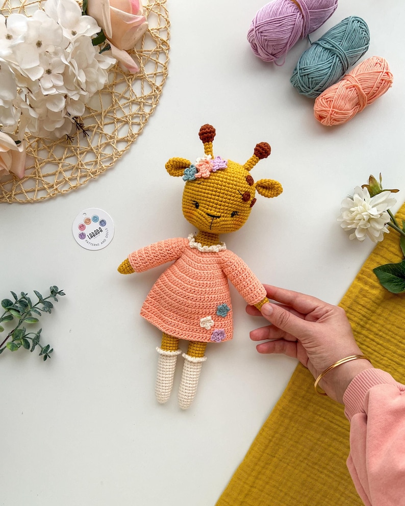 May include: A crocheted giraffe doll wearing a pink dress with white crocheted socks and a flower crown. The doll is sitting on a white surface with yarn balls and flowers in the background.