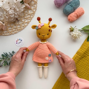 May include: A crocheted giraffe toy wearing a pink dress with white crocheted socks and a flower crown. The giraffe is sitting on a white surface with yarn balls and flowers in the background.