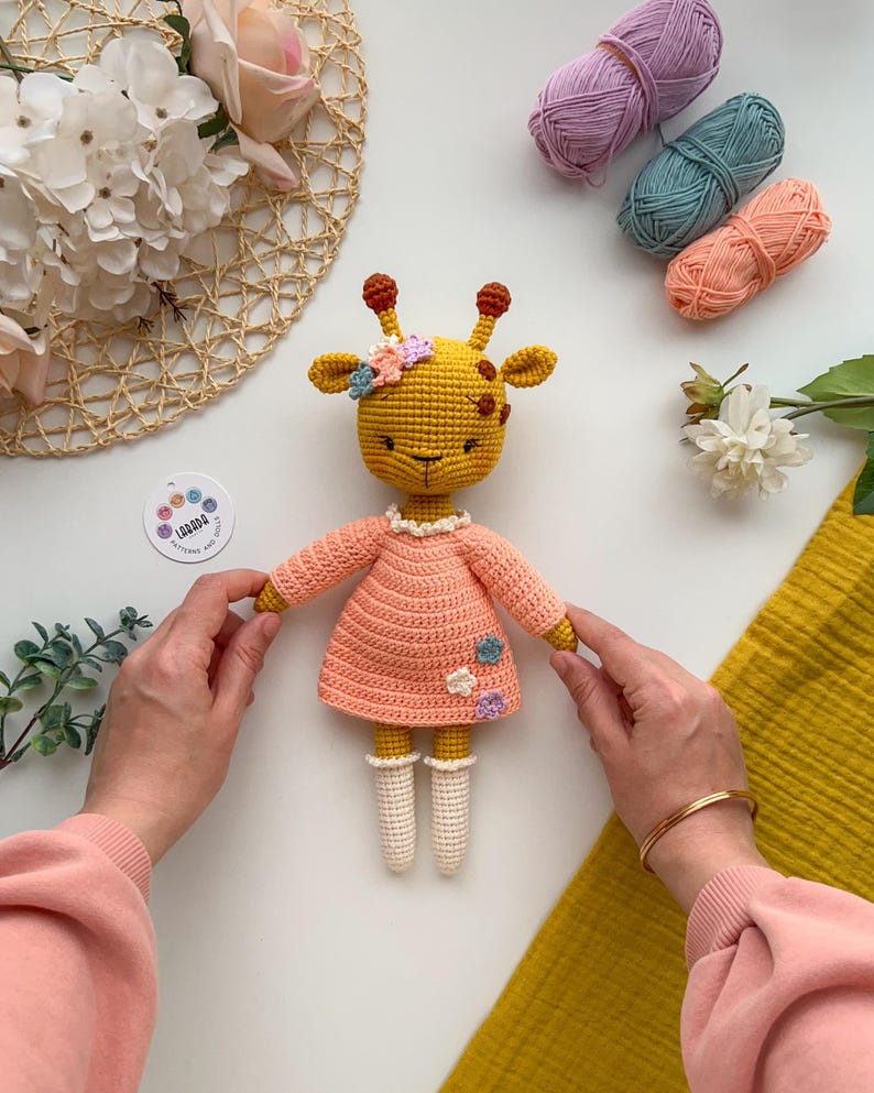 May include: A crocheted giraffe toy wearing a pink dress with white crocheted socks and a flower crown. The giraffe is sitting on a white surface with yarn balls and flowers in the background.