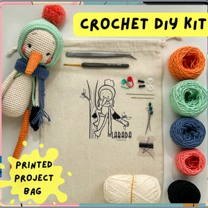 Op de afbeelding: Haak-DIY-set met een witte gehaakte vogel, garen in oranje, groen, blauw en roze, een haaknaald, naalden en een bedrukte projecttas met de tekst "Labada Crafts".
