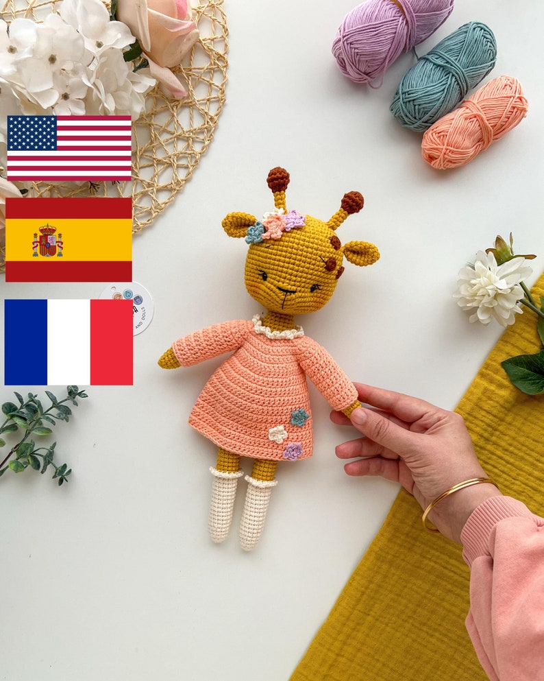 May include: A crocheted giraffe toy wearing a pink dress with white crocheted socks and a flower crown. The giraffe is sitting on a white surface with yarn balls and a woven basket in the background.