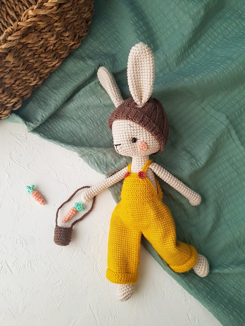 May include: A handmade crochet bunny doll wearing yellow overalls and a brown hat. The bunny has long ears, a small pink nose, and is holding a small brown bag with two crochet carrots. The doll is approximately 12 inches tall.