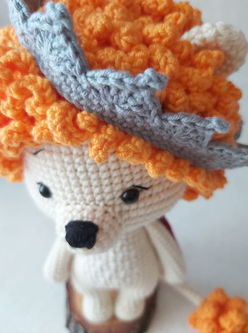 May include: A crocheted lion toy with a gray crown and orange mane. The lion is sitting on a wooden base.