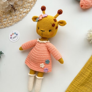May include: A crocheted giraffe doll wearing a pink dress with white crocheted socks. The giraffe has a yellow body with brown spots and a flower crown.
