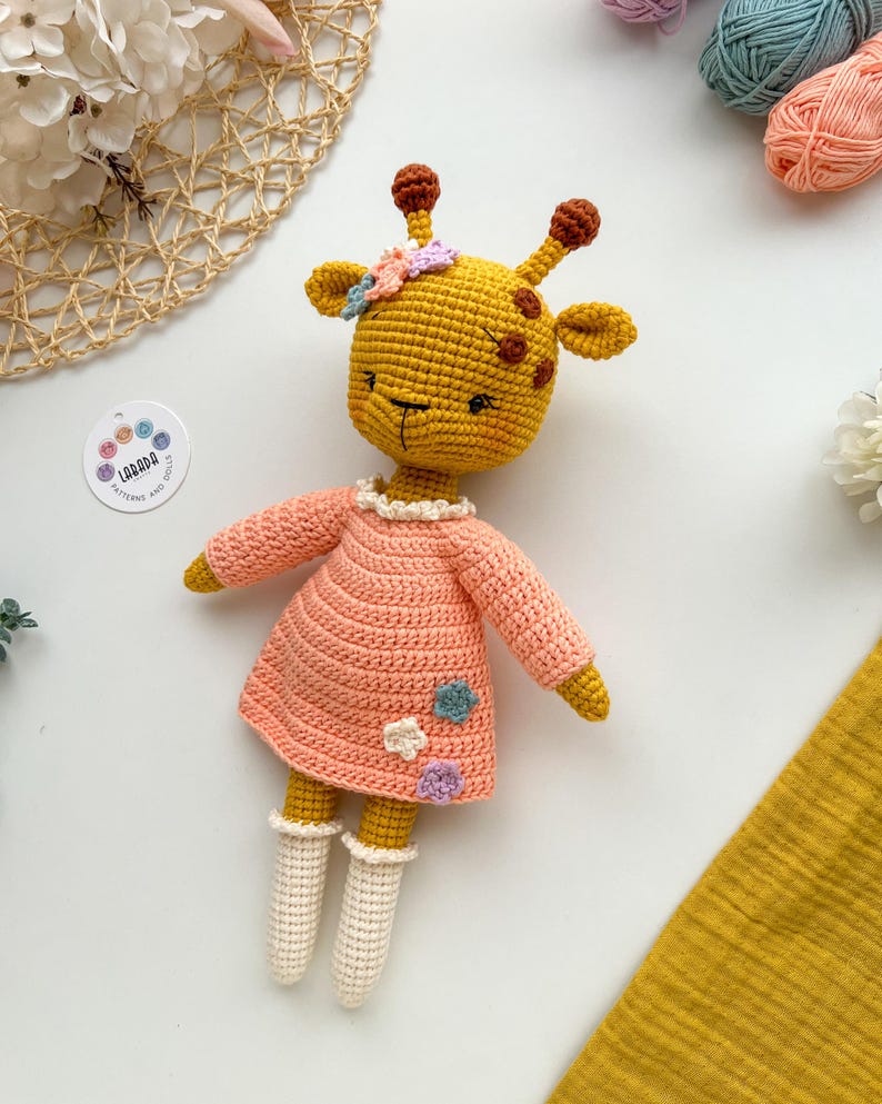 May include: A crocheted giraffe doll wearing a pink dress with white crocheted socks. The giraffe has a yellow body with brown spots and a flower crown.