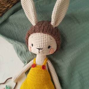 May include: A handmade crochet bunny doll with a cream-colored face and ears, wearing a brown cap and yellow overalls with red buttons. The doll has rosy cheeks and a stitched face. The background is a green fabric.
