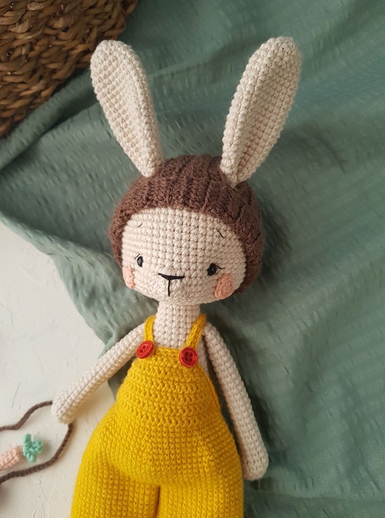 May include: A handmade crochet bunny doll with a cream-colored face and ears, wearing a brown cap and yellow overalls with red buttons. The doll has rosy cheeks and a stitched face. The background is a green fabric.