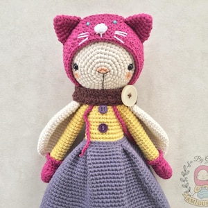 Può includere: Una bambola di coniglio all'uncinetto con un cappello da gatto rosa, un maglione giallo, una sciarpa marrone, una gonna viola e guanti rosa. La bambola ha ali bianche e un naso a bottone.
