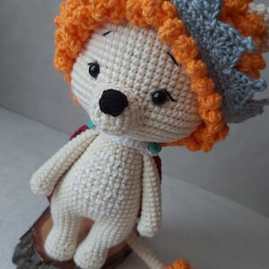 May include: A crocheted lion toy with a white body, orange mane, and a gray crown. The lion is standing on a wooden base and has a small orange tail.