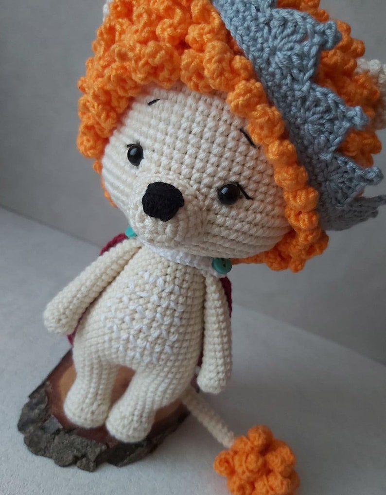 May include: A crocheted lion toy with a white body, orange mane, and a gray crown. The lion is standing on a wooden base and has a small orange tail.