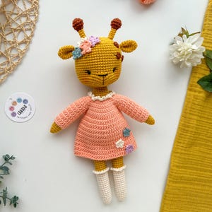 May include: A crocheted giraffe toy wearing a pink dress with white crocheted socks and a flower crown. The giraffe is light brown with brown spots and has a pink nose and black eyes.