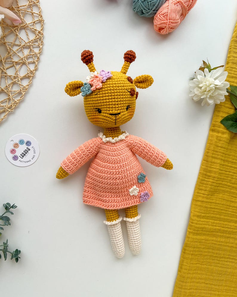 May include: A crocheted giraffe toy wearing a pink dress with white crocheted socks and a flower crown. The giraffe is light brown with brown spots and has a pink nose and black eyes.